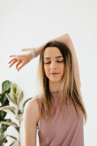 A young woman with long blonde hair, wearing a sleeveless pink top and smartwatch, stands with her eyes closed and one arm raised peacefully in a bright room.