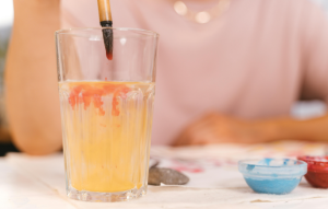 A close-up of a paintbrush being rinsed in a glass of yellow-tinted water, with red pigment dispersing, and small paint containers in the background.