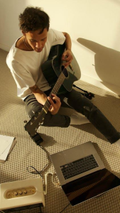 A man sitting on a couch, playing guitar in front of a laptop.