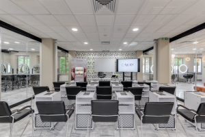 A modern training classroom with black salon chairs and white desks facing a whiteboard and digital display screen in a bright, glass-walled facility.