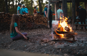 A girl in a hoodie sits alone beside a bonfire at night.