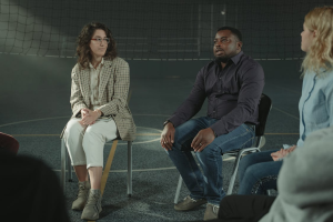 A group of people are seated on black and gray chairs in a therapy session.