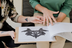 A person holding a clipboard with a black inkblot image while another person gestures with their hand.