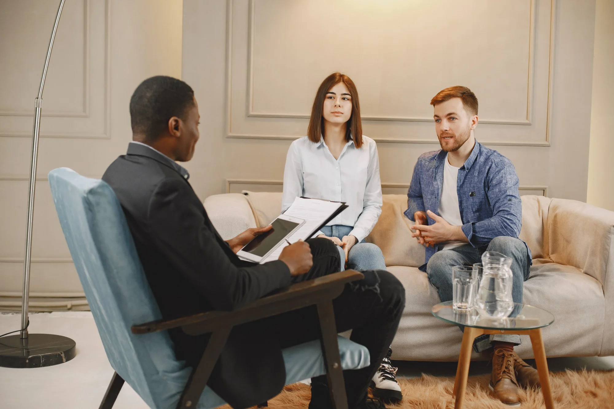 A man and woman sit on a couch, engaged in conversation with another man nearby.