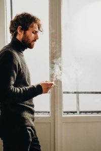 Man wearing a black sweater standing beside a window while smoking.